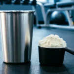 شیکر استیل و اسکوپ پودر پروتئین وی روی کف باشگاه ورزشی با نورپردازی حرفه‌ای